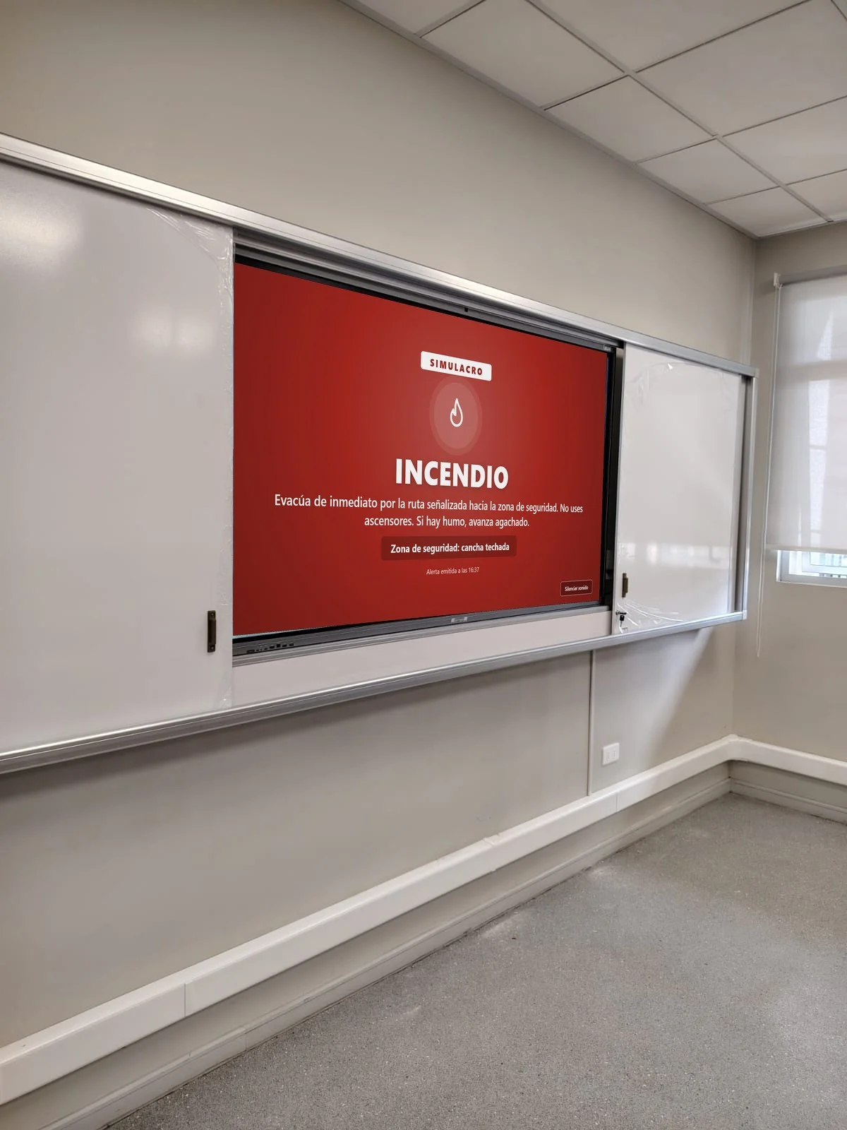 Sala de clases con la pantalla interactiva mostrando una alerta PISE de EduVoice a pantalla completa